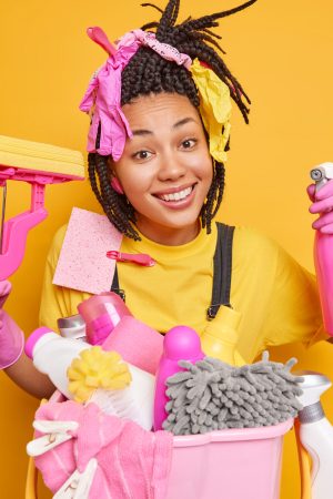 Home cleaning concept. Positive dark skinned ethnic woman with dreadlocks holds detergent and mop does laundry has cheerful expression isolated over yellow background. Housewife does house duties